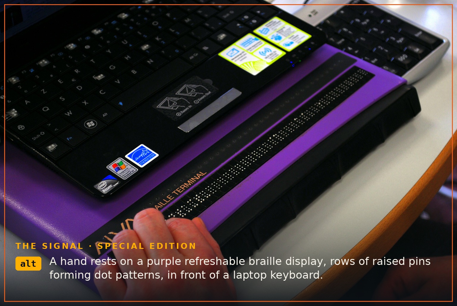 Close-up of a hand on a purple refreshable braille display in front of a laptop keyboard, with an amber caption bar displaying the photograph's own alt text.